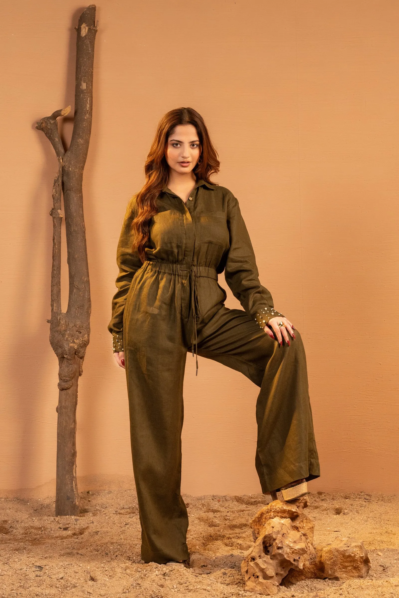 Sand-toned wall with a bare tree trunk, sand and rocks, and a model in an olive jumpsuit