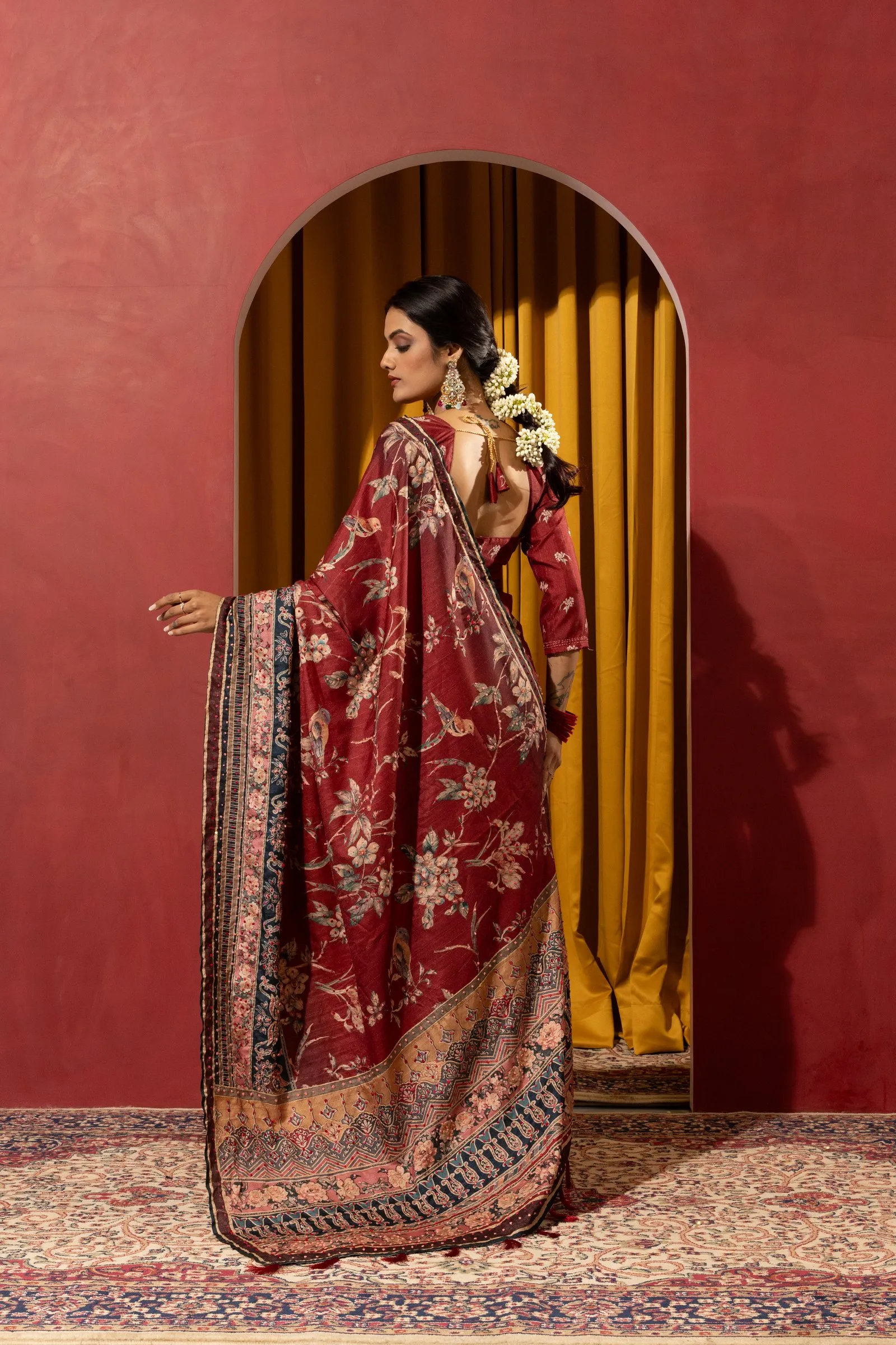 Red plaster wall with an arched doorway, mustard curtain and patterned rug, with a model in a printed saree